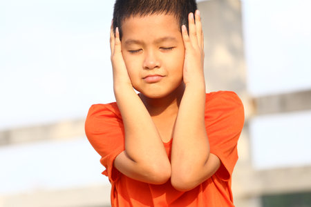 A young boy in an orange shirt covers his ears while enjoying the sunlight outdoors. His expression captures a moment of playful innocence and tranquility.の写真素材