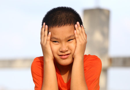A young boy in an orange shirt looks distressed while holding his hands to his face. The emotion captures a moment of worry or contemplation against a bright outdoor background.の写真素材