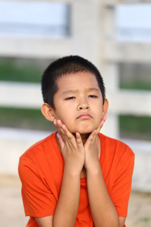 A thoughtful boy wearing an orange shirt shows a pensive expression while resting his hands on his face. Emphasizing innocence and deep contemplation.の写真素材