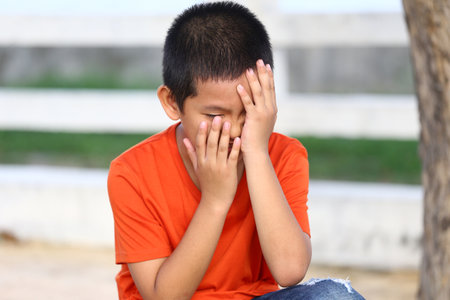 A young boy in an orange shirt sits outdoors, expressing deep emotion by hiding his face with his hands. This candid moment captures vulnerability and reflection.の写真素材