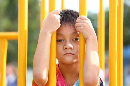 A young child leans against brightly colored playground bars, reflecting feelings of sadness and isolation amidst playtime. The image captures raw emotions in an outdoor setting.の写真素材