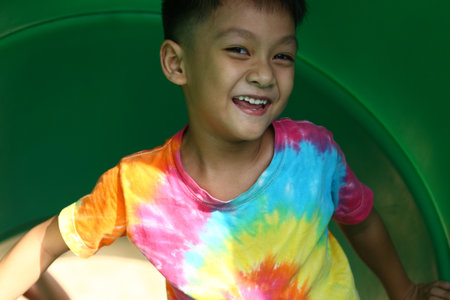 A joyful boy smiling brightly while playing on a colorful slide. His vibrant tie-dye shirt adds to the cheerful atmosphere of outdoor fun and adventure.の写真素材