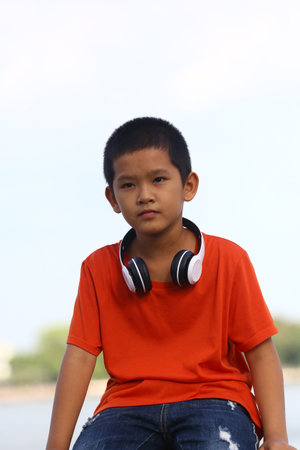 A young boy wearing headphones sits outdoors in a vibrant orange shirt. His relaxed posture and focused expression reflect a carefree and joyful vibe in nature.の写真素材