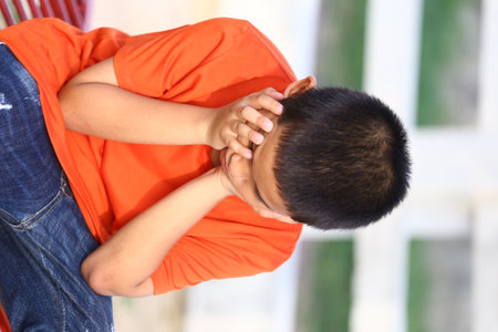 A young boy in an orange shirt sits outside, covering his face with his hands. His expression reflects a moment of deep emotion and vulnerability.の写真素材