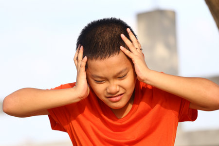 A young boy in an orange shirt demonstrates a moment of distress outdoors, showcasing raw emotions of frustration and discomfort against a bright backdrop.の写真素材