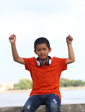 A cheerful boy in an orange shirt, wearing headphones, raises his arms in a joyful pose by the water. This vibrant scene captures youthful energy and happiness outdoors.の写真素材