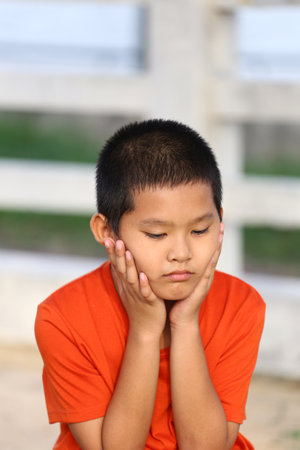 A pensive young boy in an orange shirt sits outdoors, resting his chin on his hands. His thoughtful expression conveys deep emotion and introspection.の写真素材