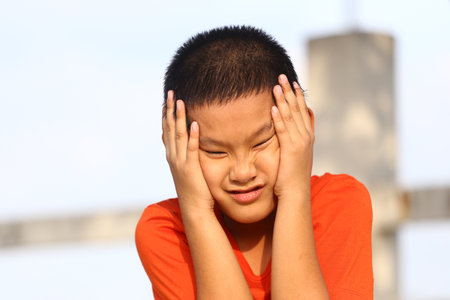 A young boy in a bright orange shirt shows frustration while holding his head outdoors, capturing genuine childhood emotion in a vibrant setting.の写真素材