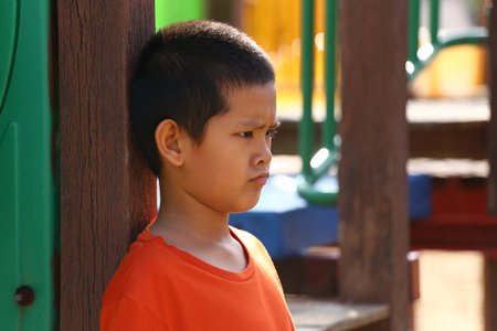 A young boy stands thoughtfully against a wooden post on a playground, wearing a bright orange shirt, showcasing a pensive expression in the sunlight.の写真素材