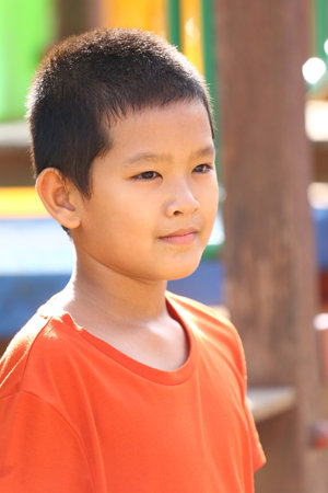 A young boy with short hair is seen standing in an outdoor playground. He wears a bright orange shirt and has a thoughtful expression as he enjoys the surroundings.の写真素材