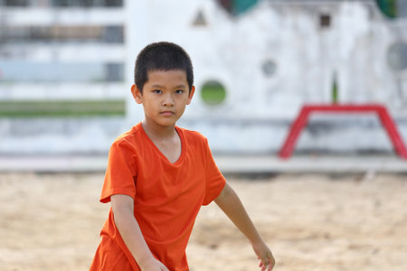 A young boy wearing an orange shirt walks playfully on the sandy ground of a playground, capturing the essence of childhood joy and innocence.の写真素材