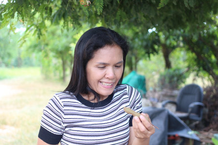 A woman smiles while enjoying a snack outdoors, surrounded by lush greenery. This candid moment captures the joy of eating in a natural setting.の写真素材