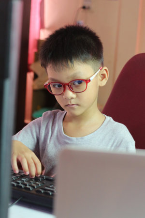 A focused young boy wearing glasses sits at a desk, engaging with a laptop. His concentration reflects the importance of learning in a digital age.の写真素材