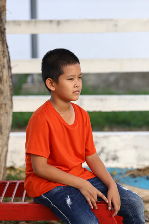A thoughtful boy sits on a red bench in a park, wearing an orange shirt and jeans. The scene captures a moment of quiet reflection and innocence in nature.の写真素材