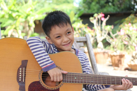 A joyful young boy sits outdoors in a garden, playing his guitar. His cheerful smile and playful stance capture the essence of childhood creativity and leisure.の写真素材