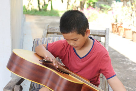 A young boy sits outdoors, concentrating on playing his guitar. The sunlight creates a warm atmosphere as he explores his musical talent in a peaceful setting.の写真素材