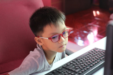 A young boy wearing glasses is intently focused on his computer screen. The scene captures the essence of curiosity and the joy of learning in a modern environment.の写真素材