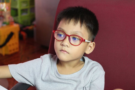 A thoughtful child wearing bright red eyeglasses sits indoors, exuding calmness and concentration. The portrait captures youthful innocence and focus.の写真素材