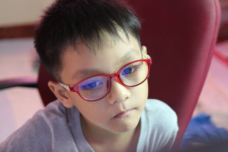 A close-up portrait of a thoughtful boy wearing red glasses, expressing a serious demeanor while sitting indoors. Perfect for themes of education and youth.の写真素材
