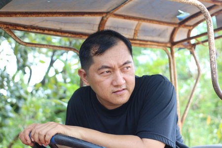 A contemplative man gazes thoughtfully while resting on a rustic vehicle amidst a lush green backdrop, capturing a moment of tranquility and reflection in nature.の写真素材