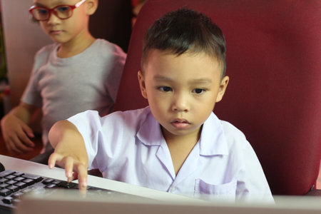 A young boy in a light-colored shirt is focused on a computer screen, exploring technology. Behind him, another child observes, highlighting a playful learning environment.の写真素材