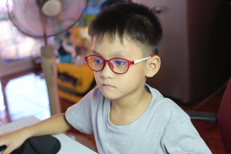 A young boy wearing red glasses sits at a desk, focused on a computer screen. The setting reflects a modern workspace, emphasizing technology and learning.の写真素材
