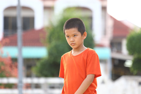 A young boy in a bright orange shirt looks thoughtfully outdoors, capturing a moment of contemplation in a serene park setting filled with greenery.の写真素材