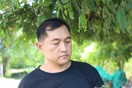 This image features a man in a black shirt reflecting thoughtfully under lush green trees, capturing a serene moment in nature on a bright day.の写真素材