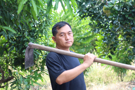 A farmer stands confidently with a tool in hand amidst lush greenery, showcasing the essence of rural life and agricultural dedication in a vibrant outdoor setting.の写真素材