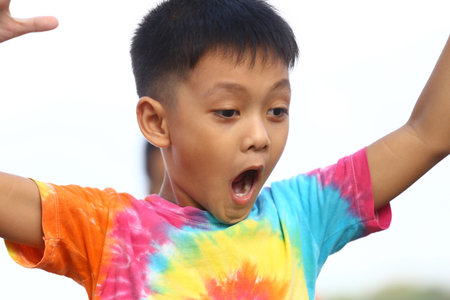 A young boy expresses sheer excitement while wearing a vibrant tie-dye shirt. Captured outdoors, his joyful reaction radiates carefree energy and innocence.の写真素材