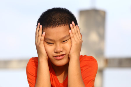 A young boy in an orange shirt shows a mix of frustration and emotion while outdoors. His hands are on his face, capturing a candid moment of expression.の写真素材