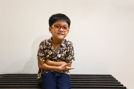 A cheerful young boy with glasses sits comfortably on a bench, exuding happiness and confidence. His casual shirt and playful pose add to the warmth of the scene.の写真素材