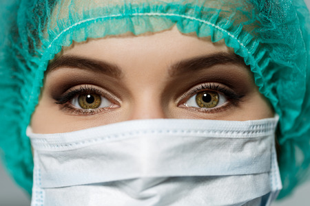 Female doctor's face wearing protective mask and green surgeon's cap closeup. Surgeon's eyes close up gazing intently in camera. Resuscitation concept.の写真素材