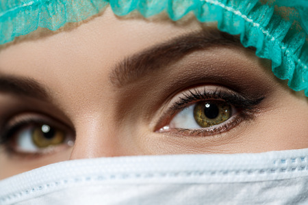 Female doctor's face wearing protective mask and green surgeon's cap closeup. Surgeon's eyes close up gazing intently in camera. Resuscitation concept.の写真素材