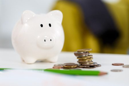 Pile of different coins near white piggybank on table closeup. Budgeting expenses concept. Making savings and effective investment concept Pile of different coins near white piggybank on table closeup. Budgeting expenses concept. Making savings and effective investment concept