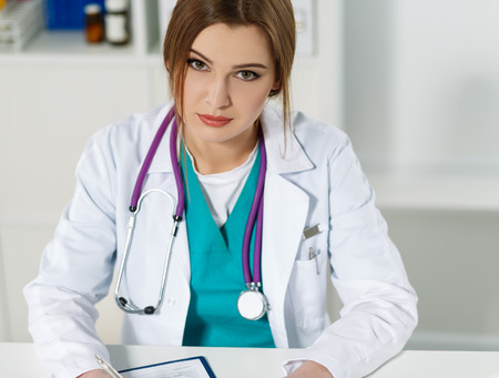 Beautiful young female therapist doctor sitting in front of working table smiling and looking in camera. Physician waiting for patient or listening carefully to him. Medical conceptの写真素材