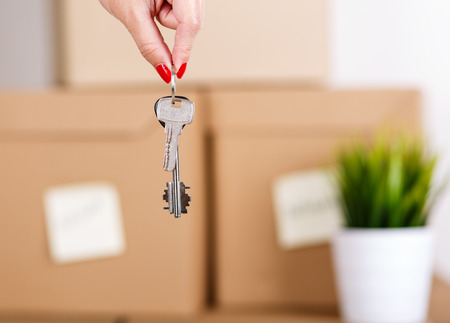 Female hand holding keys over pile of brown cardboard boxes with house or office goods background. Moving to new place of living concept.の写真素材
