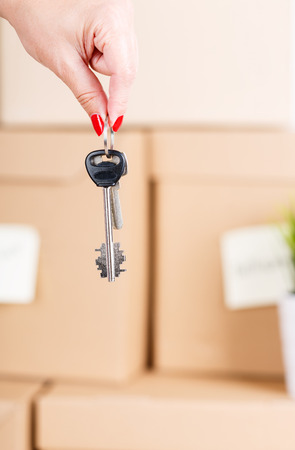 Female hand holding keys over pile of brown cardboard boxes with house or office goods background. Moving to new place of living concept.の写真素材