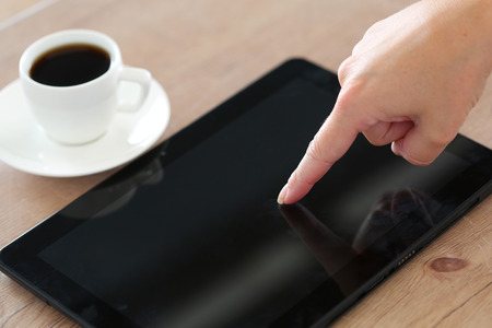 Female hand pointing in black tablet pc and using it closeup with cup of coffee in background.
