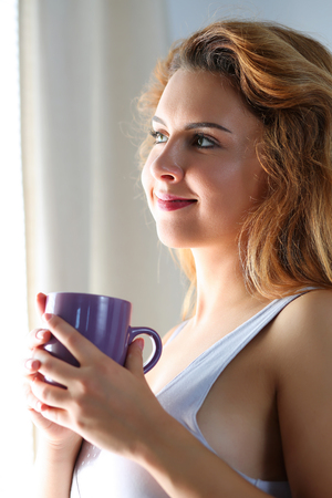 Beautiful red haired young woman looking in window and drinking morning cup of coffee or tea. New sunny day and fresh hot beverage in female hands. Weekend, summer, holidays conceptの写真素材