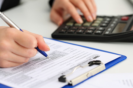 Female accountant hand holding pen counting on calculator income for tax form completion closeup.