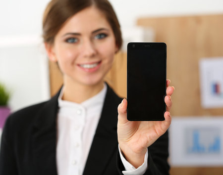 Smiling businesswoman in office hold cellphone in hands and show screen to camera. Modern communication technology, business presentation, woman in suit use smartphone searching information conceptの写真素材