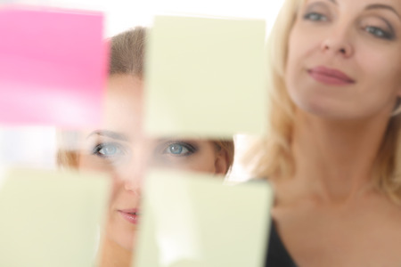 Two beautiful smiling businesswoman make some marks on sticky notes on glass wall. Fresh view, discuss problem, review situation, look at new angle, professional training, memo graph or chart conceptの写真素材
