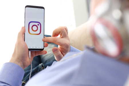Minsk, Belarus - December 25, 2018: Male arm hold samsung galaxy s9 phone with instagram social network web application logo on display closeup while lying on couch at home wearing headphonesのeditorial素材