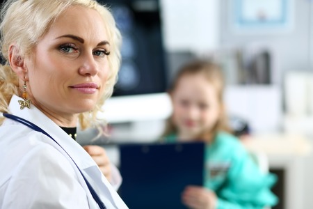 Mature blonde doctor at her office receiving little patient filling in some medical form on clipboard pad portraitの写真素材