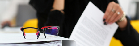 Pile of binder folders lying at worktable and stylish red black glasses closeup with female worker in background. Revision help internal revenue service agent investigation bank database workflowの写真素材