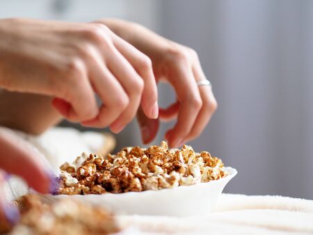 Female hands taking popcorn grains out from bowl while watching tv closeupの写真素材
