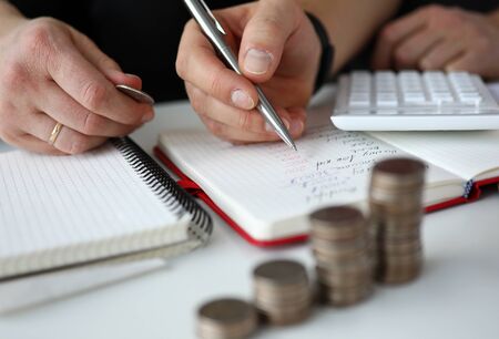 Close-up of man calculating total amount of expenses in month. Notebook with remarks. Coins and calculator on table. Family budget finance and economy conceptの写真素材