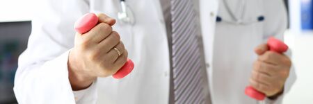 Close-up of male hands holding barbells. Physician in medical gown showing exercises with pink dumbbells to patient. Healthy lifestyle and medicine conceptの写真素材