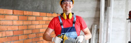 Smiling repairman in yellow hard hat holding in hands huge professional drill portrait